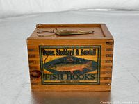 Wooden box with dovetailed corners and colorful fish hook advertisement label, fishing lure placed on top.