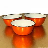 Front angle showing three orange enamel nesting bowls with white interiors lined up on a reflective surface.