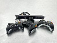 Three black Xbox 360 wireless controllers and one Kinect sensor placed together, showing device shapes and condition.