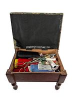 Open sewing stool showing interior storage compartment with assorted sewing supplies including black thread spool, scissors, measuring tape, needles, and other small notions.