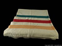 Folded cream wool blanket showing horizontal stripes in teal, red, and mustard yellow