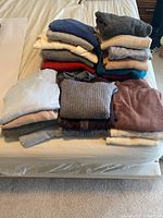 Stack of 23 folded sweaters in assorted colors on bed