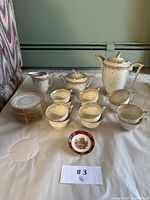 Full tea set laid out on a white sheet includes teapot, sugar bowl, cream server, 12 teacups, 12 saucers, and small wall hanging plate in front.
