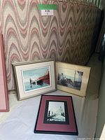 Shows three framed art prints leaning against a patterned couch: harbor scene, fishing scene, and cityscape.