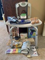 View of entire lot on small plastic table showing all items including scale, curling iron, brushes, soaps, candle, plunger, massage rollers.