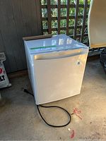 Danby white mini bar fridge front view with power cord coiled next to it on ground, located in garage environment with lattice wood behind.