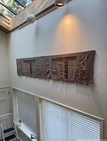 Wide shot of the large rectangular textile wall hanging with geometric pattern on a high wall in an entryway, showing the natural light from windows above and nearby blinds below.