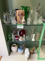Photo showing multiple clear, colored, and patterned glass vases and pitchers on two glass shelves, alongside pottery and milk glass pieces below.