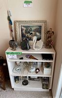 Full view of white shelf with framed art, glass swan figurines, Axicon fairy statue, wooden figurine, elephant figurine, and small decorative items inside and on top of shelves