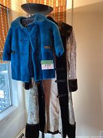 Two vintage faux fur coats hanging on a hat stand near a window. The shorter blue coat overlaps the longer tan and dark brown coat.