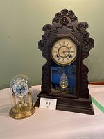 Photo of the two clocks side by side on a table: a large carved wooden mantel clock with glass front, and a smaller solar glass dome clock with brass base.