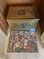 Photo showing front and top embossed surfaces of the gold tin box with intricate family scene and textured border patterns. A small wall hanging tapestry featuring a medieval horseback knight and attendants is placed folded beneath the box.