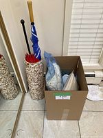 Photo of cardboard box containing scarves, hats, and gloves packed inside plastic bags; umbrellas visible behind box in umbrella stands