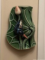Front view of green ceramic wall pocket vase shaped as a large lotus leaf with detailed veining and textured stem, featuring a sculpted bird figure in blue, white, and orange perched on the leaf's raised stem.