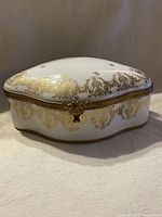 View of the entire antique porcelain trinket box showing the white porcelain body with intricate gold leaf hand painting and the bronze-colored metal trim around the edges.