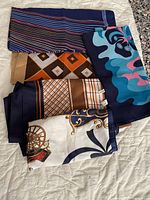 Five scarves laid out on a white surface showing various patterns including stripes, abstract colors, geometric and carriage motifs
