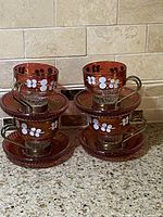 Four vintage cranberry glass espresso and macchiato cups with metal holders stacked on matching saucers, showing white hand painted floral patterns.