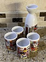 Photo showing the porcelain sake decanter with narrow neck and flared rim along with four cylindrical sake glasses, all decorated with colorful Maneki Neko lucky cat illustrations.
