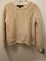 Full view of vintage ivory cable knit sweater hanging on hanger showing traditional textured patterns across body and sleeves.