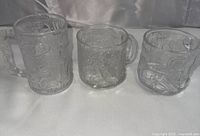 Photo of three clear 3D glass mugs showing embossed Batman Forever character designs and sculpted handles, set on a white drape.