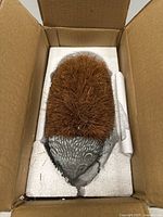 Hedgehog-shaped brush packed in a box with styrofoam protection, bristles brown and dense, base detailed with hedgehog face design in grey plastic.