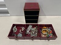 Top view of red jewelry box with three open drawers filled with assorted jewelry pieces.