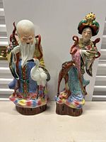 Two detailed porcelain figurines side by side: elderly man with staff and crane, woman with fan and dog