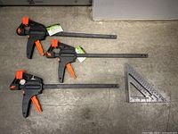 Three ratchet bar clamps laid flat on a concrete floor with a triangular carpenter's square for scale.