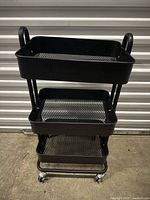 Black metal rolling storage cart with three mesh trays and caster wheels against a closed white garage door.