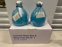 Two blue glass wasp and bee traps with white netting tops and hanging strings displayed on a white box labeled 'Colorful Glass Bee & Wasp Trap Set Of 2'.