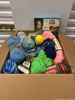 Top down view of a cardboard box filled with various colored wool yarn balls and skeins, with a box of electric scissors on top.