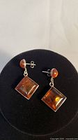 Pair of drop earrings with square amber stones and round amber studs on a black display stand, showing front view.