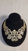 Front view of gold coloured antique style necklace displayed on black bust, showing ornate floral and scroll motifs