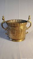 Full front view of the brass and copper urn with two handles, showing hand-tooled imagery.