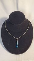 Full view of sterling silver chain necklace with blue glass teardrop pendant displayed on black stand.