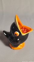 Front left angle view of black ceramic crow matchstick holder showing large open orange beak containing matchsticks and orange feet.