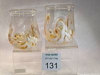 Two handblown Kingston Glass Studio "Twisty Cups" with golden and white swirl design on clear glass displayed on acrylic blocks.