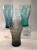 Three Coca-Cola glasses on clear acrylic stands showing front and side profiles documenting color, embossing, and base details.