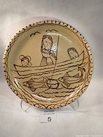 Full view of ceramic pottery plate with hand-painted folk art scene of man and children in boat with fish and birds, shown standing on clear display stand on white background.