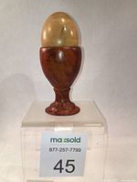 Egg cup holding the wooden egg displayed on a clear acrylic stand with lot label visible.