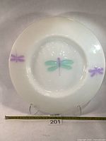 Full front photo of large white milk glass charger with a central green and purple dragonfly and two purple dragonflies near the rim, on stand with measuring tape showing size.