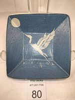 Full top view of blue square pottery dish showing white heron with raised wings, water rushes, and full moon design in corner.