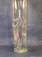 Front view of the tall clear cylindrical glass showing embossed dancing nude female figures design wrapped around the glass.