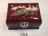 Front view of red lacquered jewelry music box with detailed mother of pearl and seashell peacock and floral inlay on lid, showing glossy finish and metallic snap clasp.