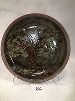 Full view of circular stoneware plate with dark glaze and reddish-brown tree motif in center on display stand.