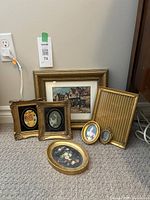 Seven small gold tone framed art pieces arranged on carpeted floor, showing variety in shapes, sizes, and art styles including portraits and a village scene.