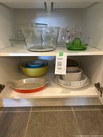 Two shelves of clear Pyrex glass bakeware and bowls, including mixing bowls, baking dishes, measuring cups, and a green glass lemon squeezer.