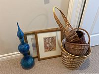 Overall lot showing six woven baskets stacked/nested, two framed landscape prints leaning against wall, and a tall blue-green ceramic accent vase.