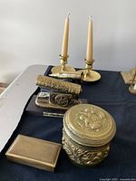 Full view of the lot on a table with a pair of brass candlestick holders with candles, brass cannon shaped stapler, brass cannon shaped lighter, and round brass decorative box.