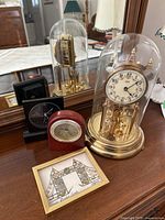 Photo of the Kundo gold tone mantel clock under glass dome, black quartz clock, wooden Michael C Fina quartz clock, and framed Tower Bridge art on a dresser with mirror